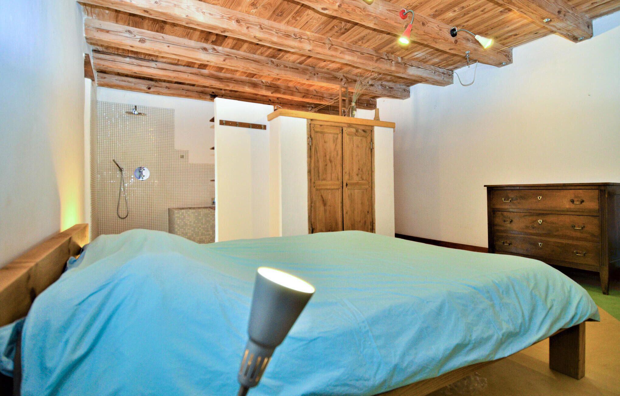 Une chambre avec un plafond en bois, un lit recouvert d’une courtepointe bleue, une commode en bois et une armoire en bois.