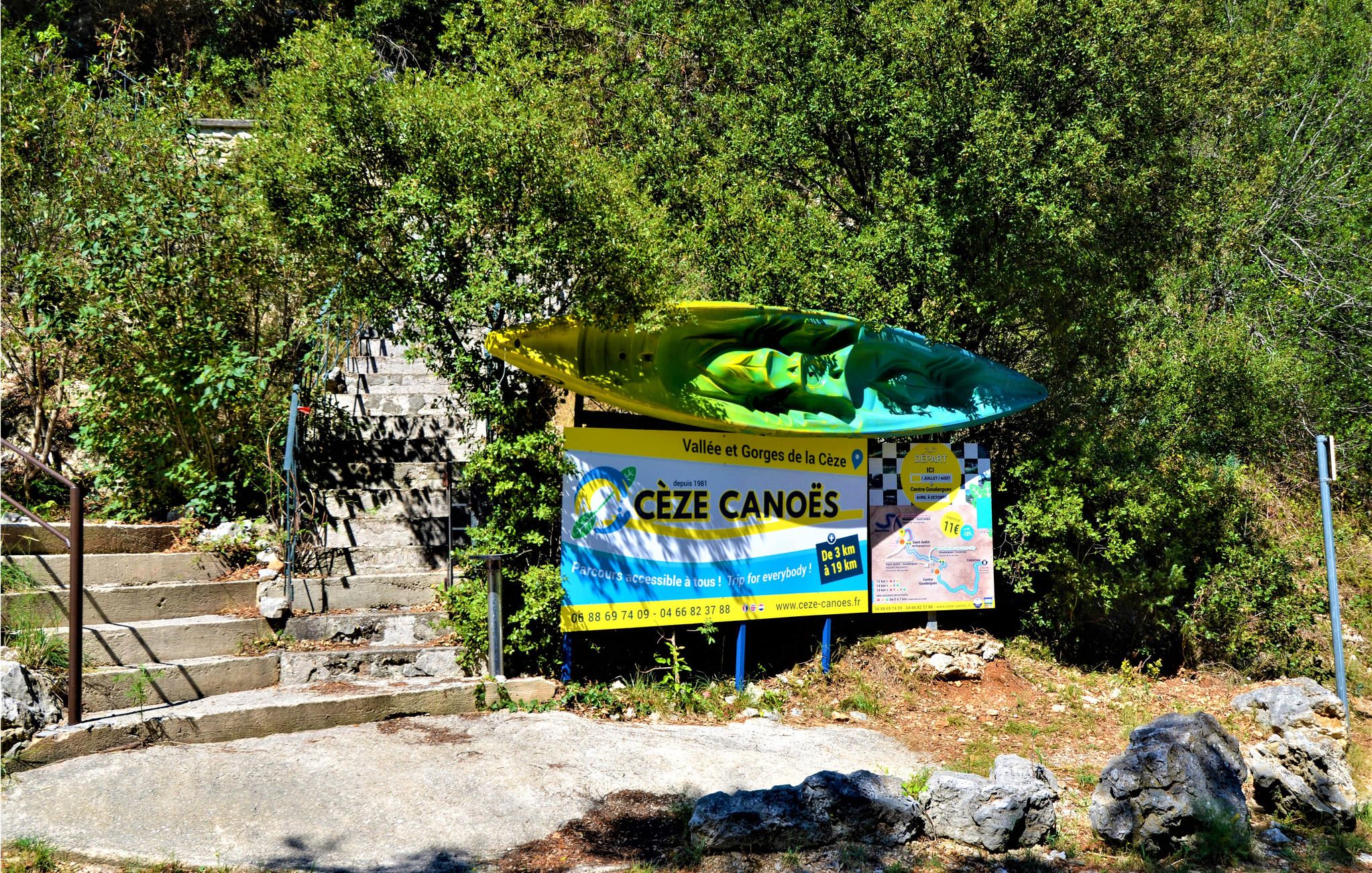 A sign for Cèze Canoës with a kayak and a map, located near a staircase and surrounded by greenery.