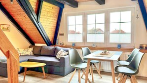 A cozy loft-style room with a sloped ceiling, a sofa, a dining table with chairs, and large windows offering a view of the outside.