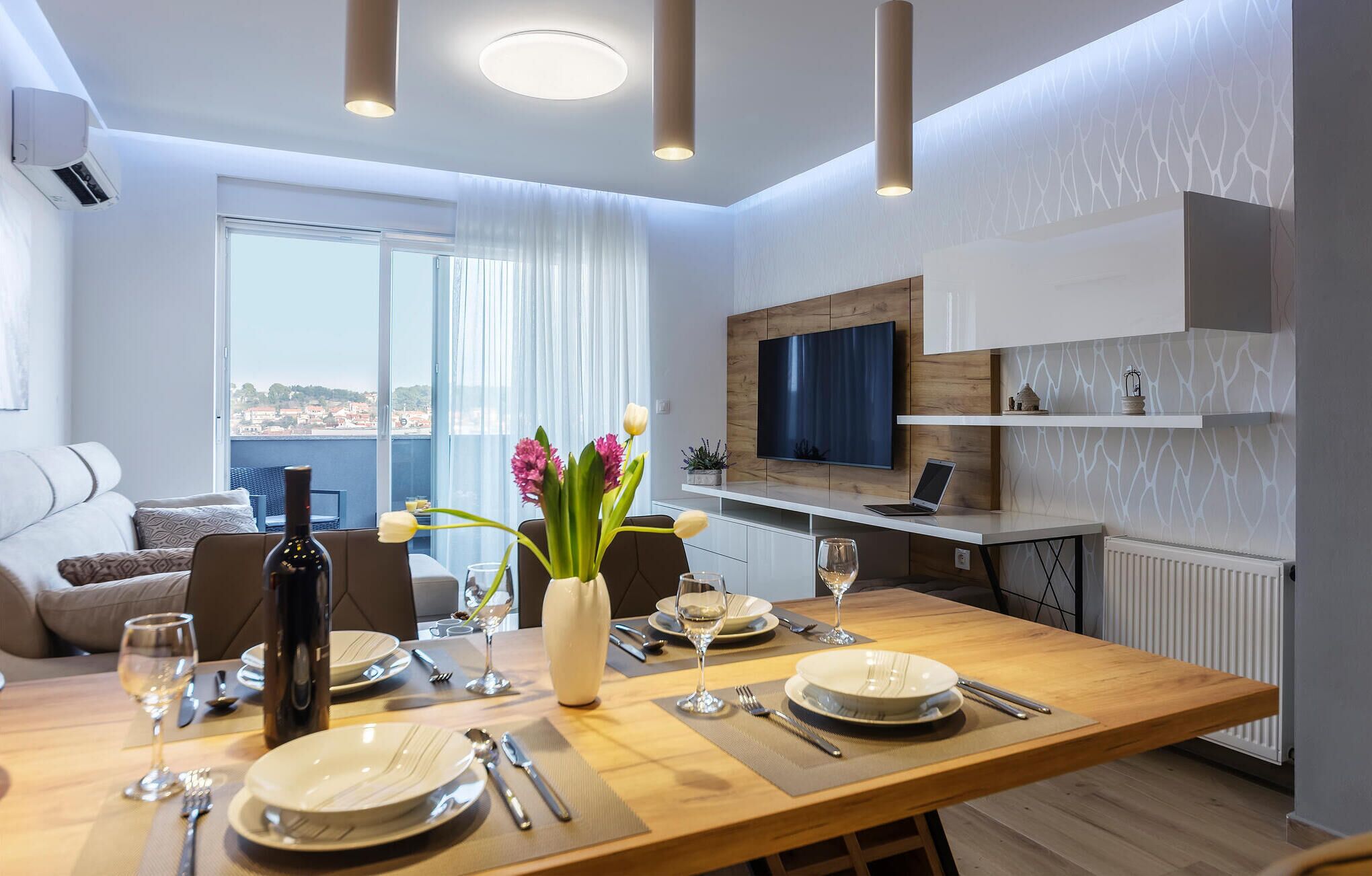 A modern dining area with a wooden table set for four, a large window with curtains, a flat-screen TV mounted on the wall, and a view of the city.