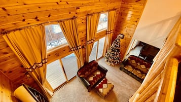 A cozy cabin interior with a wooden ceiling, large windows with curtains, a decorated Christmas tree, and a fireplace.