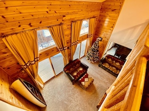 Interior acogedor de una cabaña con techo de madera, ventanas amplias con cortinas, un árbol de Navidad decorado y una chimenea.