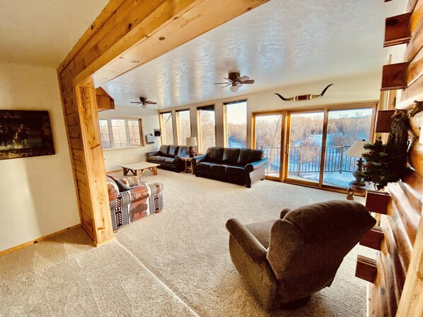 A spacious living room with a fireplace, large windows, and a ceiling fan.