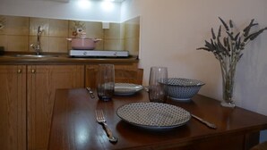 Una mesa de comedor de madera con un plato, un vaso y un tenedor. Se observa una cocina al fondo con una estufa y armarios.