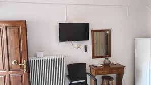 Una habitación con puerta de madera, un radiador, una silla negra, un escritorio de madera con un jarrón, un espejo y un televisor de pantalla plana montado en la pared.