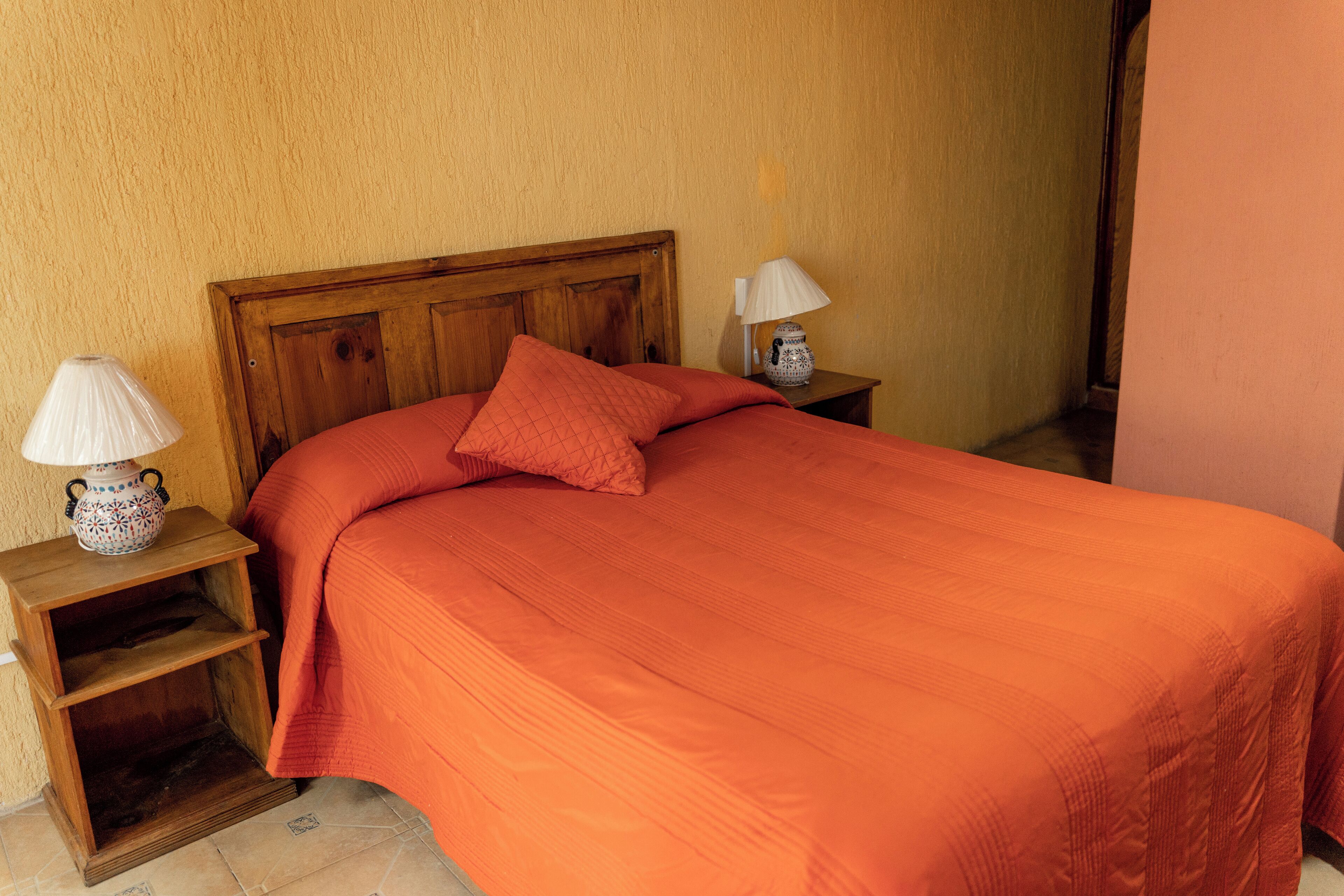 A bedroom with a wooden headboard, a single bed with an orange bedspread, two bedside tables with lamps, and a small shelf.