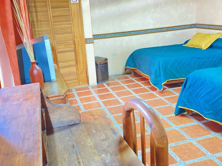 Una habitación con dos camas, un escritorio de madera, una silla, un cesto de basura pequeño y un armario de madera.