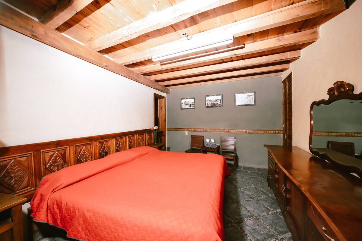 Une pièce avec un plafond en bois, un lit recouvert d’une couverture rouge, une commode, un miroir et des tableaux encadrés accrochés au mur.