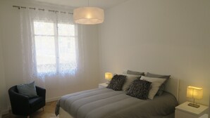 Un dormitorio con una cama, dos mesitas de noche, una silla, una ventana con cortinas y una lámpara colgante.