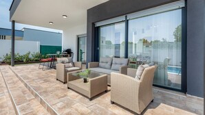 An outdoor patio with wicker furniture, a glass table, and a view of a swimming pool through large windows. - Beach front home in Peroj (Peroj)