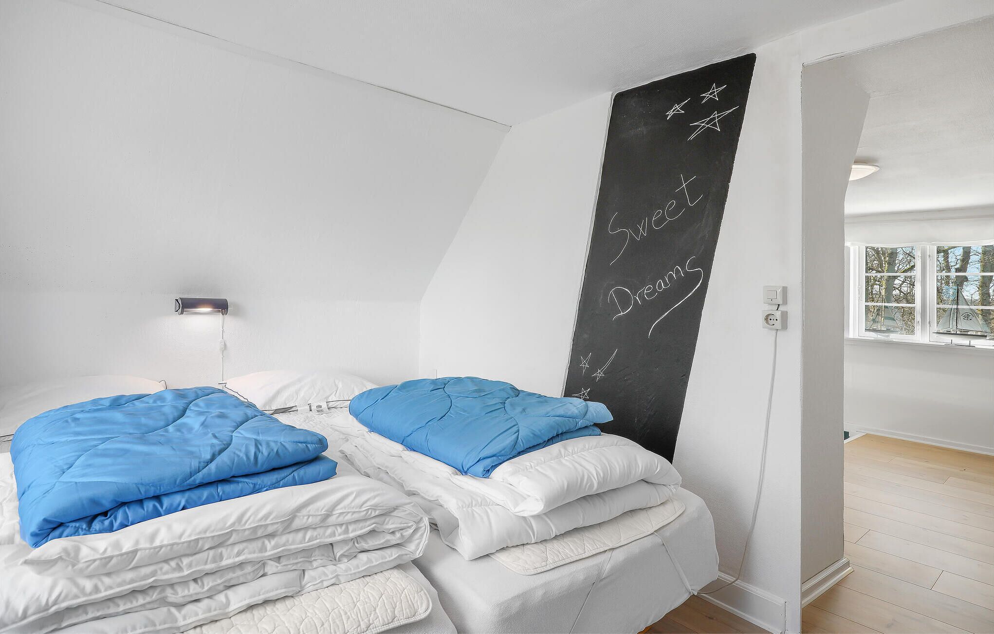 Un dormitorio con dos camas, una pizarra con escritura, una lámpara de noche y una ventana con un modelo de velero.