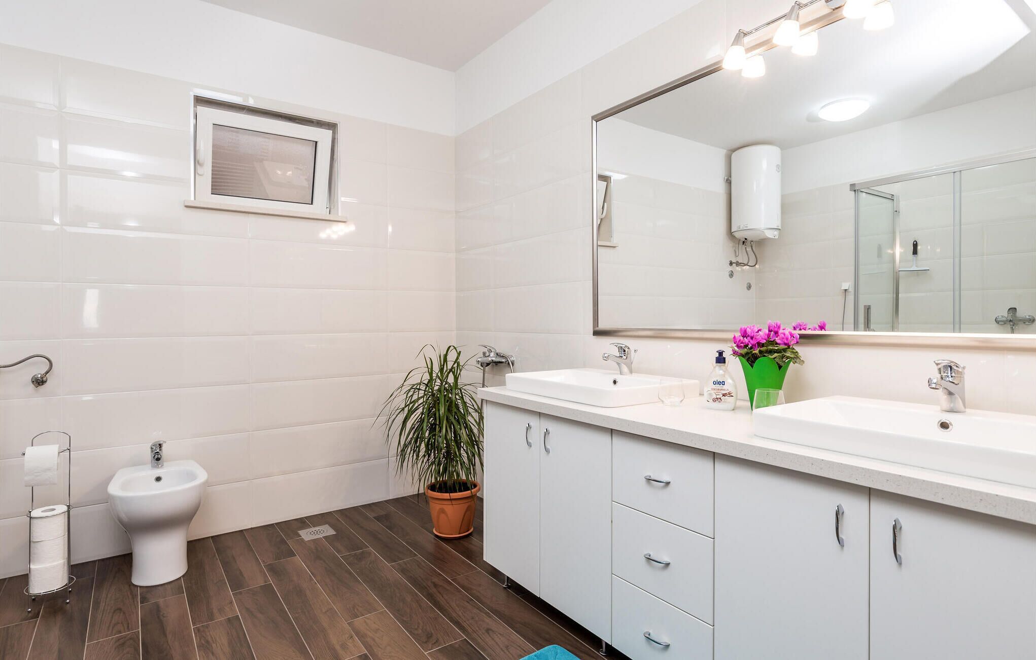 A modern bathroom with white tiles, a bidet, a sink with a mirror, and a potted plant.
