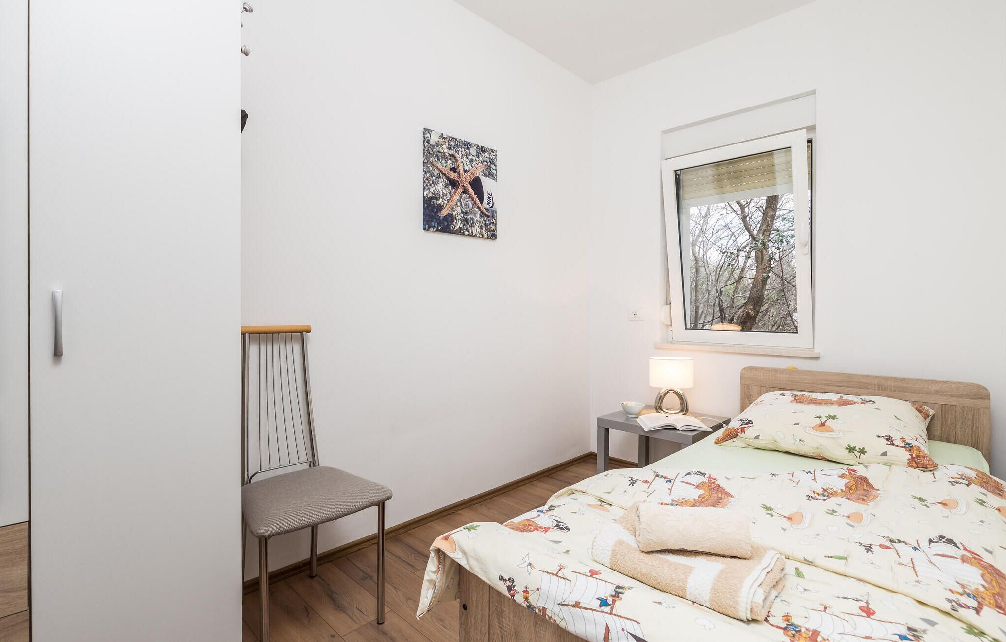 Een slaapkamer met een bed, een stoel, een nachtkastje, een raam met uitzicht op bomen en een muur met een ingelijste foto.