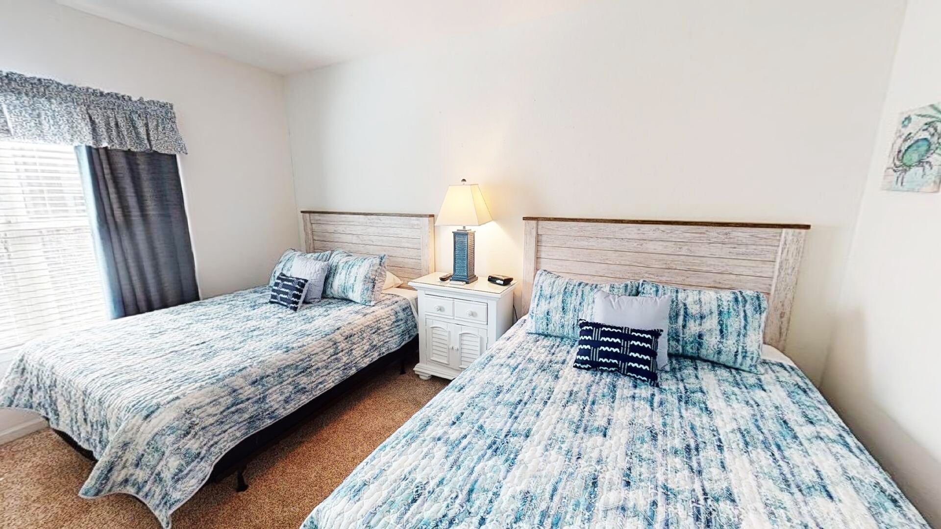 A room with two beds, each with matching bedding and pillows, a bedside table with a lamp, and a window with curtains.