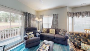 A living room with a grey sofa, armchair, and patterned armchair, a coffee table, and large windows with curtains. - Light and bright 1st floor condo in Sunset Beach | RC 1802 (Sunset Beach)
