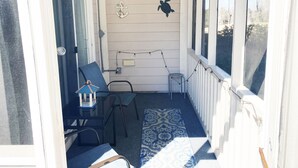 A covered porch with a blue patterned rug, two chairs, a small table, and decorative wall art including an anchor and a turtle. - Light and bright 1st floor condo in Sunset Beach | RC 1802 (Sunset Beach)