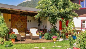 Cómodo espacio exterior con sillas de mimbre y una terraza de madera, rodeado de abundante follaje y plantas en macetas.