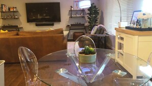 A modern living room with a glass dining table, a brown sofa, a TV mounted on the wall, and a lamp on a side table. - 2 Master Bedrooms, Luxury beds, Pool & Hot Tub, Large TVs, Surround Sound (Scottsdale)