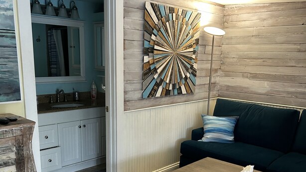 Ein Wohnzimmer mit einem Holztisch, einem dunkelgrünen Sofa, einem großen abstrakten Wandbild und einem durchgehenden Türrahmen sichtbaren Küchenbereich.