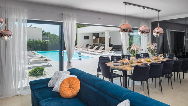 A modern dining area with a pool view, a blue sofa, and a table set for dining. - Beautiful home in Sivati with sauna (Sivati)
