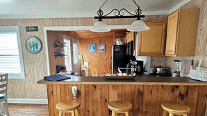 A kitchen with wooden paneling, a bar area with stools, a clock, and various appliances. - Shore Beats Work! (2040 Sq ft) (Kill Devil Hills)