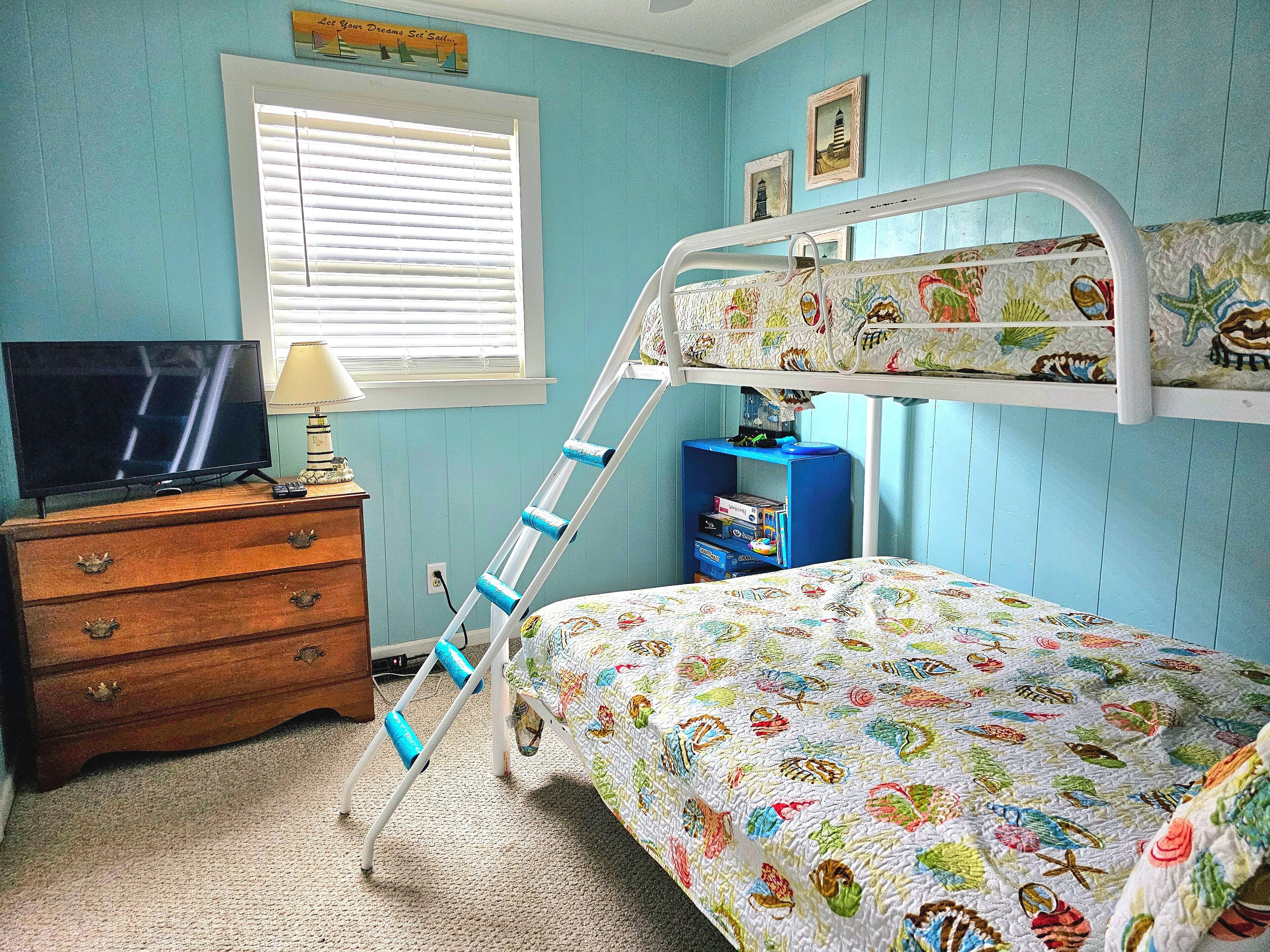Un lit superposé avec une couverture aux motifs colorés représentant des poissons, une commode en bois avec des tiroirs, un téléviseur posé sur le dessus, et une fenêtre avec des stores.
