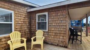 A wooden deck with two Adirondack chairs, a table, and a covered area with a playground in the background. - Shore Beats Work! (2040 Sq ft) (Kill Devil Hills)