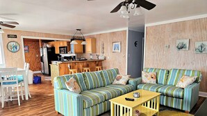 A living room with striped sofas, a wooden coffee table, and a kitchen in the background. - Shore Beats Work! (2040 Sq ft) (Kill Devil Hills)