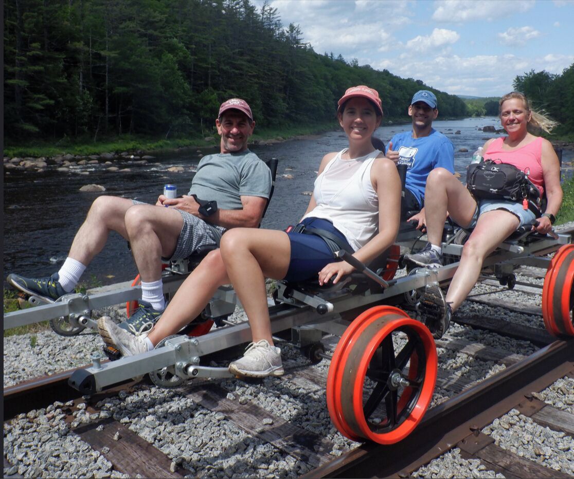 Four people sitting on a train carriage with a river and forest in the background.