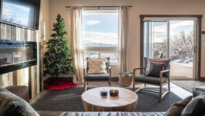 Un salon chaleureux avec une grande télévision, un sapin de Noël, deux fauteuils, une table basse et une vue sur des montagnes enneigées par de grandes fenêtres.