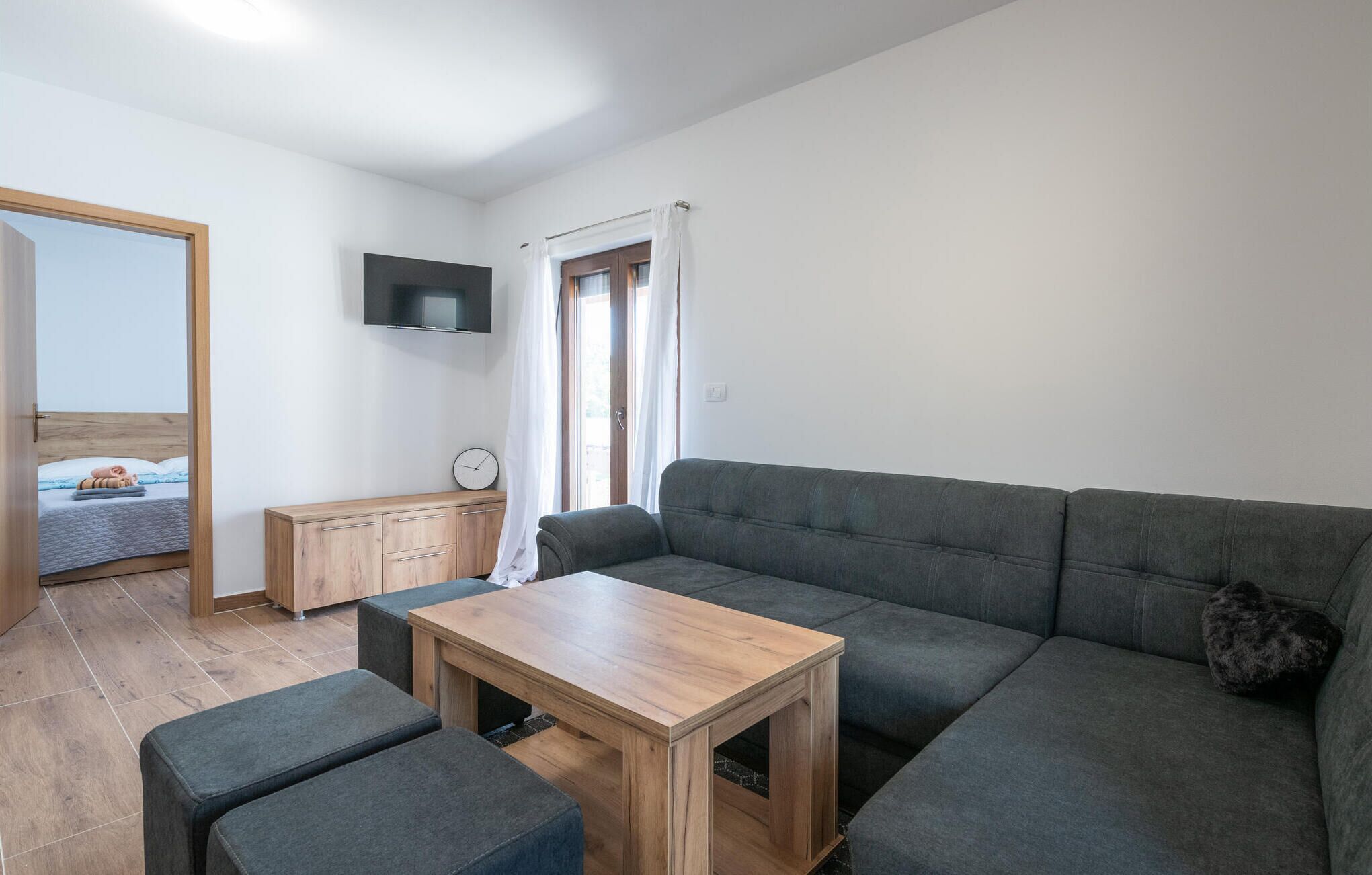 A modern living room with a grey sofa, a wooden coffee table, and a grey armchair. There is a wooden cabinet with drawers and a flat-screen TV mounted on the wall. A window with white curtains is visible, and a door leads to another room.