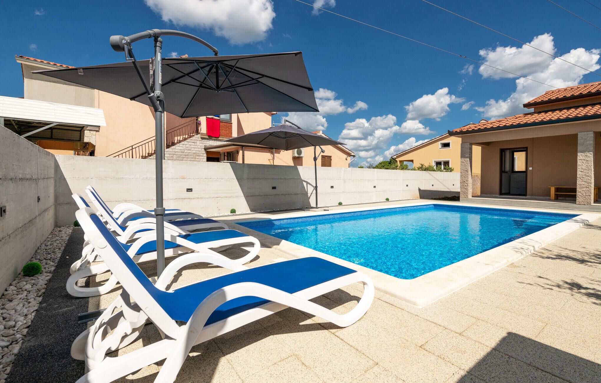 Une piscine entourée de chaises longues bleues et blanches, un espace abrité avec des parasols, et un ciel d’un bleu limpide.