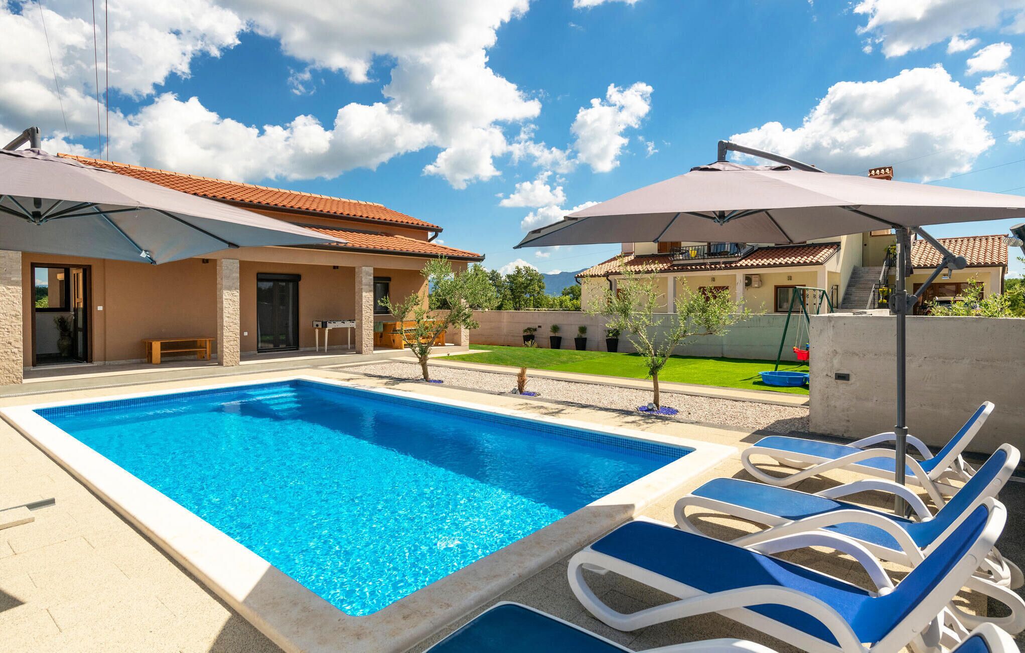 A swimming pool with lounge chairs and umbrellas, surrounded by a house with a tiled roof and a well-maintained lawn.