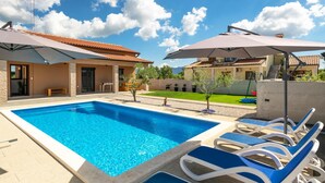 Une piscine entourée de chaises longues et de parasols, elle-même bordée d’une maison avec un toit en tuiles et une pelouse bien entretenue.