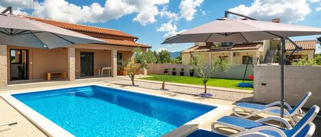 A swimming pool with lounge chairs and umbrellas, surrounded by a house with a tiled roof and a well-maintained lawn.