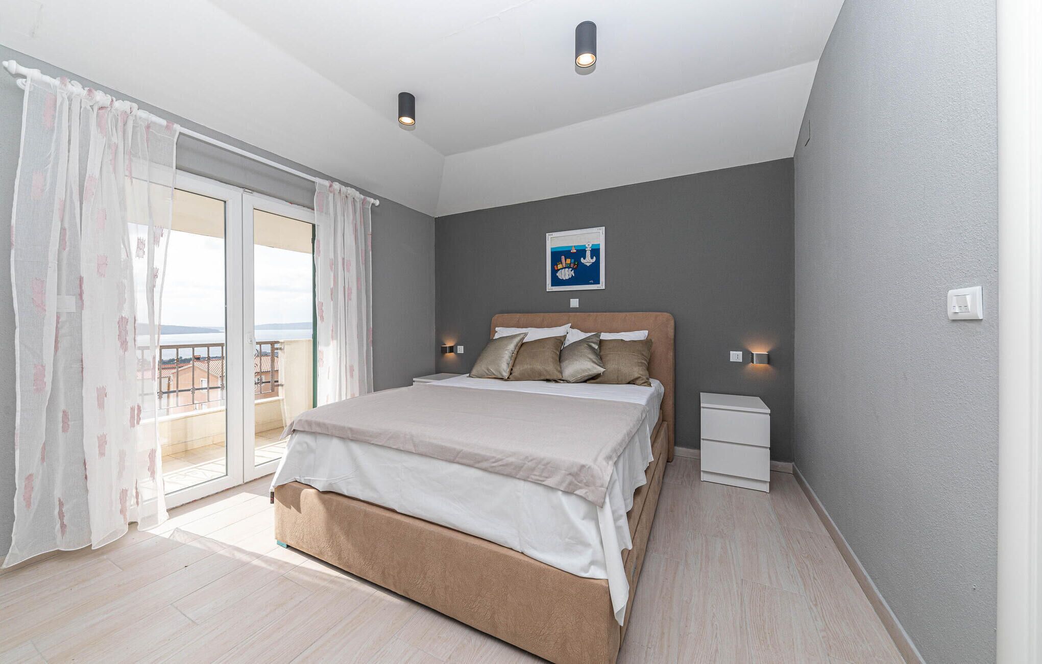 Une chambre à coucher comprenant un lit, une table de chevet, une lampe fixée au mur, une fenêtre avec des rideaux légers et une vue sur l’extérieur.