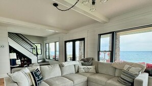 Un salon spacieux avec un grand canapé gris en forme de L, une table basse ronde en bois et une vue sur la mer grâce à de grandes fenêtres.