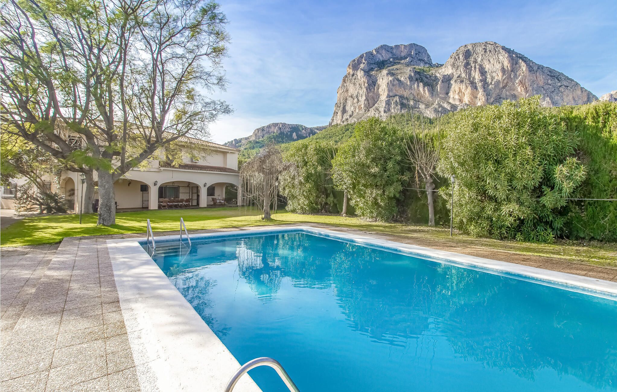 A swimming pool with a mountain backdrop, surrounded by lush greenery and a building with arches.