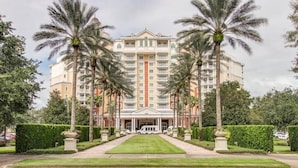 Hotel de vários andares com entrada central, rodeado por palmeiras e jardins bem cuidados.