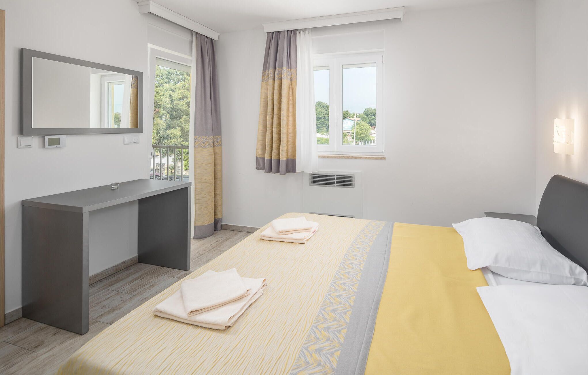 A hotel room with a bed, a bedside table, a mirror, a window with curtains, and a view of greenery outside.