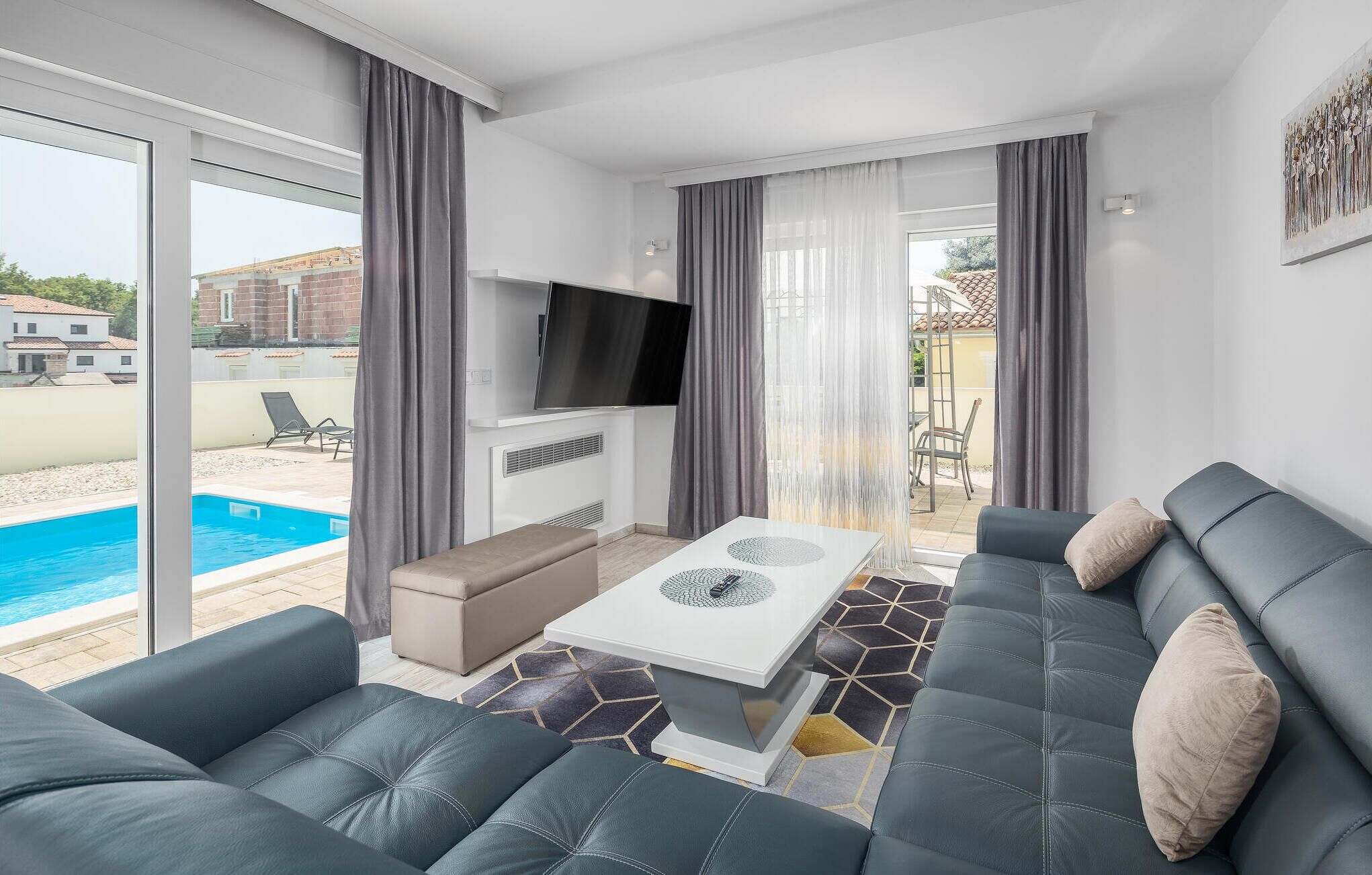 Ein modernes Wohnzimmer mit Blick auf einen Pool, grauem Sofa, einem Flachbildfernseher und einem Essbereich mit einem gemusterten Teppich.