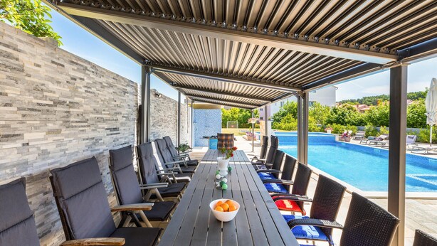 Un área de estar al aire libre techada con vista a la piscina, que incluye una mesa larga con sillas y un cuenco de fruta.