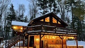 Una cabaña de madera con balcón y porche cubierto, rodeada de nieve y árboles.