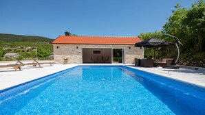 Une piscine avec un bâtiment en pierre en arrière-plan, un espace de repos couvert et une vue sur le paysage environnant.