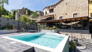 Una piscina con sillas de descanso, un edificio de piedra con balcón y vista a una estructura histórica de piedra al fondo.