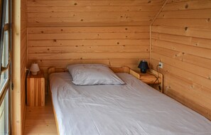 Une petite pièce de style cabane, avec des murs en bois, comprenant un lit simple, une table de chevet, une lampe et une fenêtre.