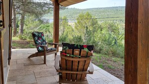 Une véranda en bois avec deux chaises et une table, offrant une vue panoramique sur un paysage verdoyant, avec des arbres et une étendue d’eau.