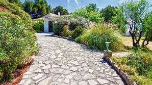 Un chemin de pierre mène à un bâtiment doté d’un toit en tuiles, qui est entouré d’une végétation luxuriante et d’arbres.