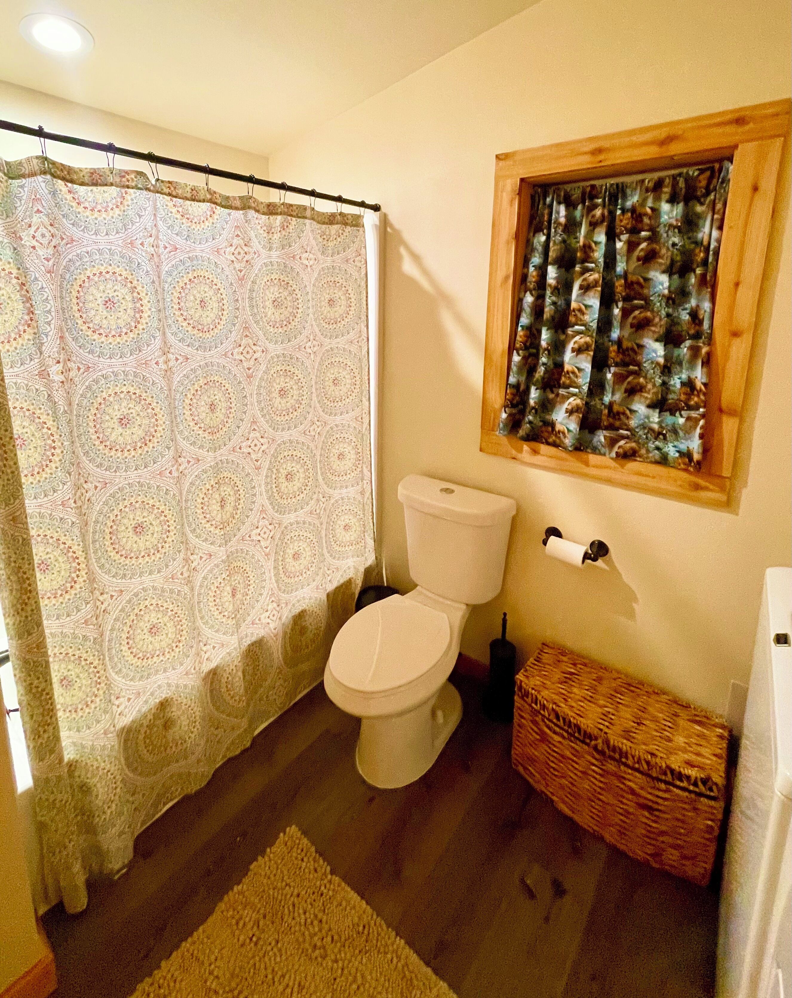 Un baño con cortina de ducha estampada, un inodoro, un ventanal con cortina y una cesta de mimbre.