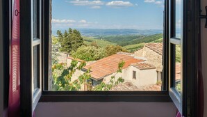 Vista da janela de uma paisagem rural com colinas ondulantes, céu claro e edifícios de pedra tradicionais com telhados de terracota.